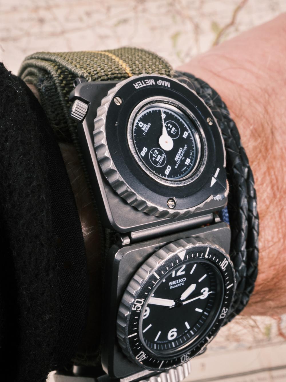 Wrist shot of a Seiko Fieldmaster Contra showing the stacked quartz time module and map meter module on a green fabric strap, highlighting the hinged modular case design against a blurred map background.