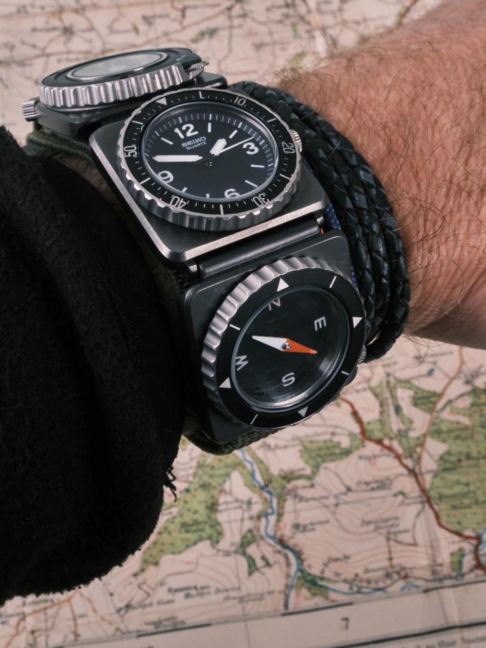 A Seiko modular field watch worn on the wrist, featuring a black quartz time module with bold Arabic numerals paired with a compass module below, both housed in square black cases on a green fabric strap, photographed against a detailed vintage map background.