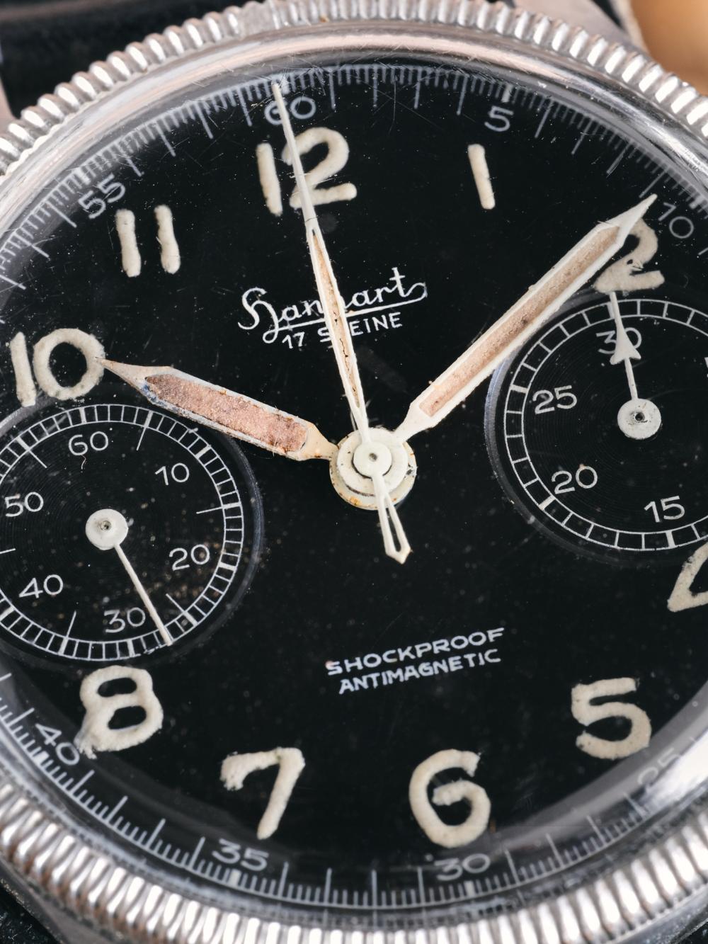 A macro close-up of the dial of a vintage Hanhart 417 ES chronograph. The black dial features aged luminous Arabic numerals, patinated syringe hands, two recessed sub-dials and the Hanhart logo beneath the 12 o'clock marker. Fine scratches on the acrylic crystal and the warm patina of the lume highlight the watch's decades of use, while the fluted stainless steel bezel frames the composition.
