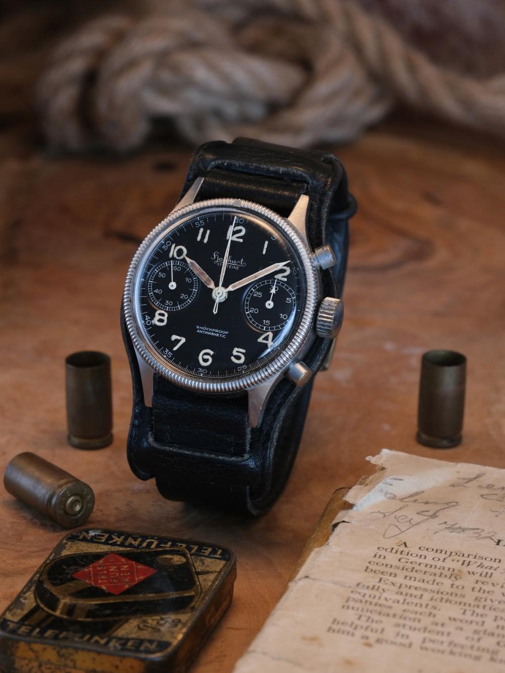 A vintage Hanhart 417 ES flyback chronograph displayed upright on its original black leather Bund strap. The stainless steel case features a coin-edge rotating bezel, black bi-compax dial, aged luminous Arabic numerals and patinated syringe hands. Period details including brass cartridge cases, a worn Telefunken tin, an aged document and coiled rope create an atmospheric setting that reflects the watch’s military heritage.
