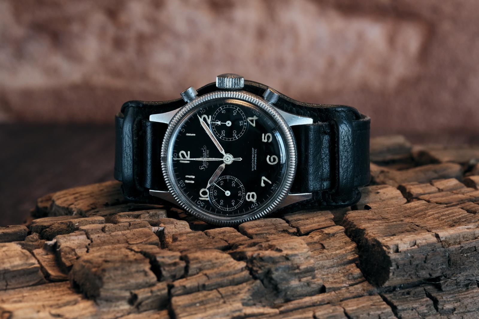 A vintage Hanhart 417 ES chronograph resting on a weathered piece of cracked timber. The stainless steel case features a fluted coin-edge bezel and a black bi-compax dial with aged luminous Arabic numerals and patinated syringe hands. Fitted with its original black leather Bund strap, the watch is photographed against a softly blurred rustic background, emphasising its military heritage and decades of honest wear.
