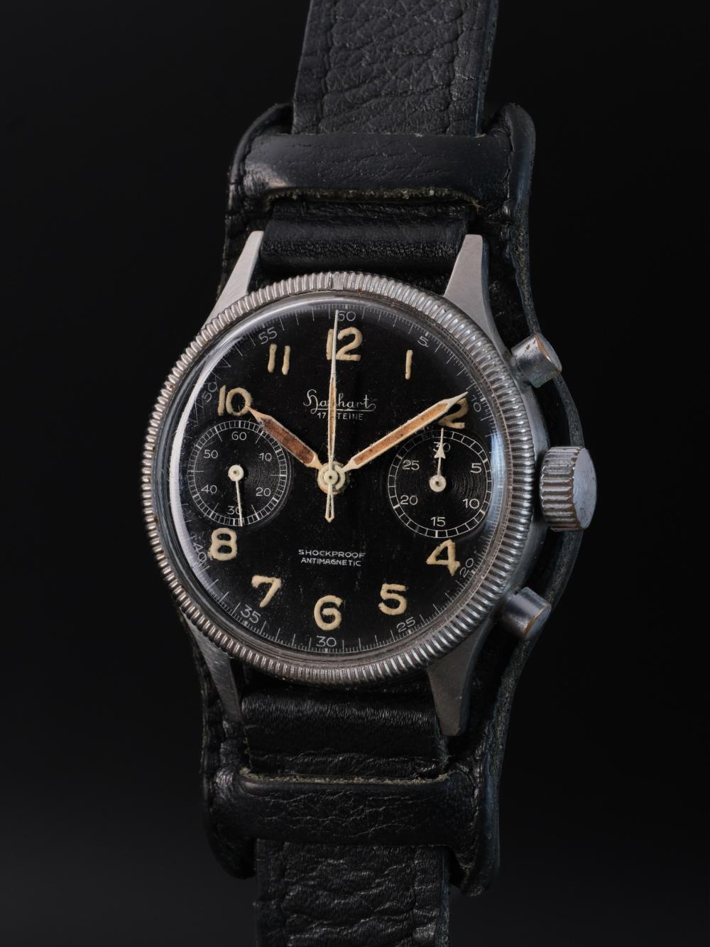 Studio portrait of a vintage Hanhart 417 ES flyback chronograph on its original black leather Bund strap. The stainless steel case features a coin-edge rotating bezel, black bi-compax dial, aged luminous Arabic numerals and heavily patinated syringe hands. Set against a dark background, the close-up highlights the watch’s honest wear, military character and beautifully preserved vintage details.
