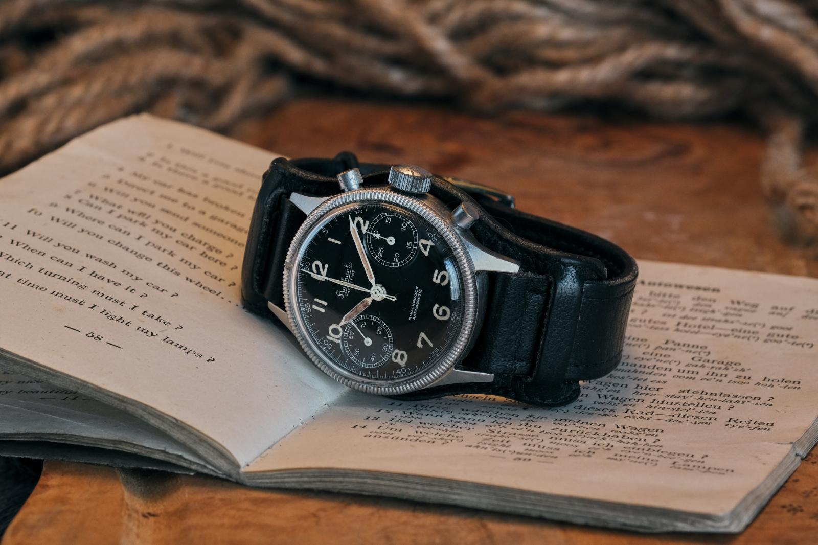 A vintage Hanhart 417 ES chronograph resting on an open period handbook. The stainless steel watch, fitted with its original black leather Bund strap, features a black bi-compax dial with aged luminous Arabic numerals, syringe hands and a fluted rotating bezel. The warm wooden surface, weathered book and coiled rope in the background create an evocative setting that reflects the watch's mid-century military heritage.
