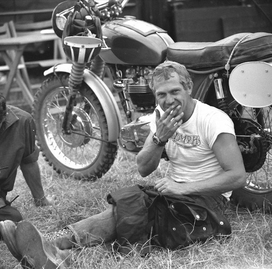 Black-and-white photograph of Steve McQueen sitting on the grass beside a vintage Triumph motorcycle, wearing a Hanhart 417 ES  chronograph on his left wrist. Dressed in a Triumph T-shirt and motorcycle trousers, he holds a cigarette while resting beside the bike, capturing the relaxed atmosphere of mid-century motorcycle culture.