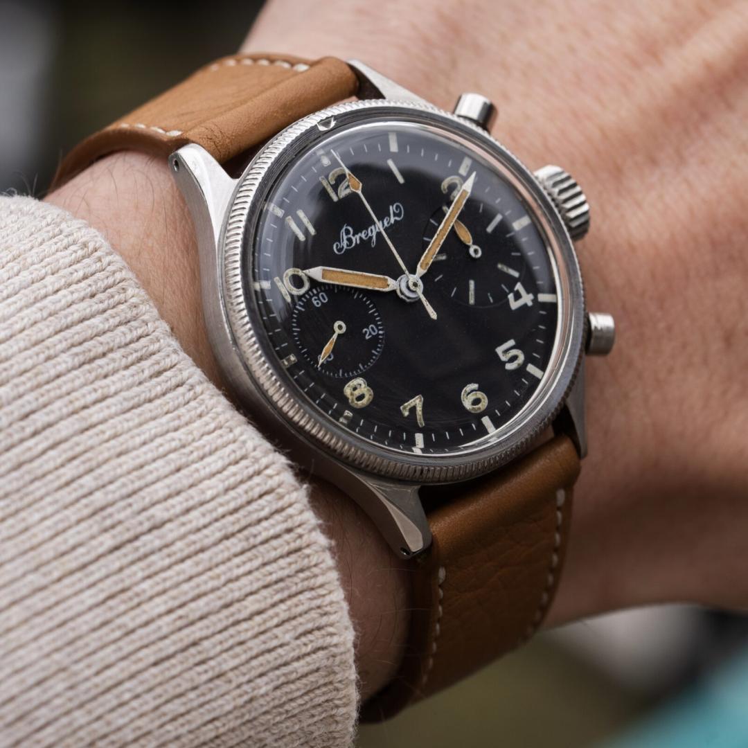 A vintage Breguet Type 20 flyback chronograph worn on the wrist with a tan leather strap. The stainless steel case features a fluted rotating bezel and black dial with aged luminous Arabic numerals, patinated hands and two chronograph sub-dials. The close-up highlights the Type 20’s functional military aviation design and distinctive vintage character.