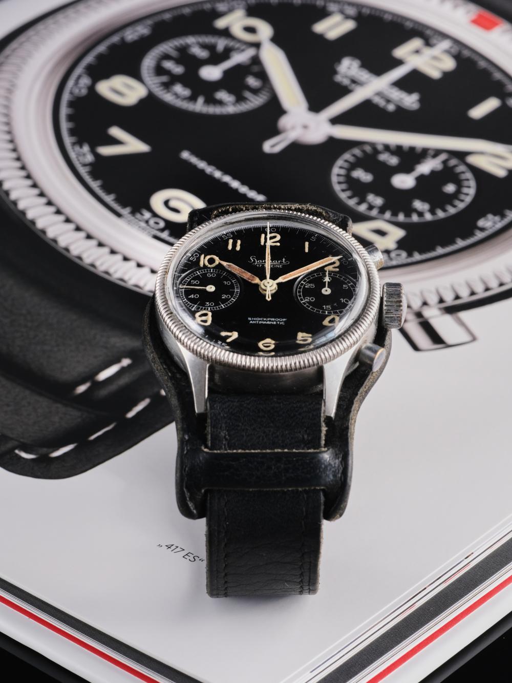 A vintage Hanhart 417 ES flyback chronograph displayed on its original black leather Bund strap, positioned over a photograph of a modern Hanhart chronograph. The composition highlights the family resemblance between past and present, from the black bi-compax dial and luminous Arabic numerals to the distinctive coin-edge bezel and military-inspired design.