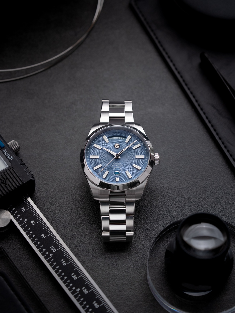 Jack Mason is a watch brand so American I wouldn’t be surprised if one of their watches got up, put on a cowboy hat and a pair of spurs and jumped into the nearest rodeo. However, while you might expect their watches to be as big and bold as a southern barbeque – and some of them are – they’re no strangers to more restrained and elegant design too. Case in point, the new Canton Day-Date, a daily wear timepiece with a classy calendar display and guilloché finish.
The Canton was previously introduced as a time and date model, meaning the primary update here is the introduction of the days of the week display at 12 o’clock as well as new colours and a new dial texture. The days function sits in a semi-circular arc between the 11 and 1 o’clock hour markers with the days written out in full. It’s a format made iconic by the Rolex Day-Date and you can definitely feel that influence here, although Jack Mason avoids too much comparison thanks to the 6 o’clock position of the date with a magnification lens below the sapphire crystal rather than above it – giving the display a markedly different layout.
Plus, where Rolex’s Day-Date predominantly has a sunray brushed finish across its face, the Jack Mason Canton Day-Date uses guilloché. Guilloché is a style of engraving that creates dramatic geometric patterns like the cross-spiral of diamonds seen here. It’s a technique traditionally performed by hand but in modern watchmaking the term is also used for milled engraving by equipment like CNC machines. For a modern brand like Jack Mason founded in 2015, the latter is the most practical option.
There are three colours of dial available: Slate, Lagoon and Chocolate. Slate is a cold, grey-blue tone that reminds me of ice while Lagoon is much more of a teal/green-blue like tropical waters. Chocolate is pretty self-explanatory in brown, though it does feature some extra accents absent on the other models such as the gold-coloured hands and hour markers.
Housing this updated dial is a 41mm diameter steel case with a 12mm thickness. It’s built for everyday wear with 150m water resistance protecting it from the weather as well as any accidental spills or pool-side related incidents. It has a smooth bezel and brushed lugs that lead into a three-link steel bracelet fitted with micro-adjustment clasp.
Inside, and visible through the exhibition caseback, is the Sellita SW240-1 top grade movement. It has a 40-hour power reserve and an accuracy of +/- 5 seconds per day (regulated in the USA). It’s fitted with a custom gold-coloured rotor with a curving profile inspired by the horns of longhorn cattle – did I mention they’re a Texan brand? That rotor is set against a backdrop of fine finishing that includes perlage, blued screws and brushing. 
Jack Mason Canton Day-Date